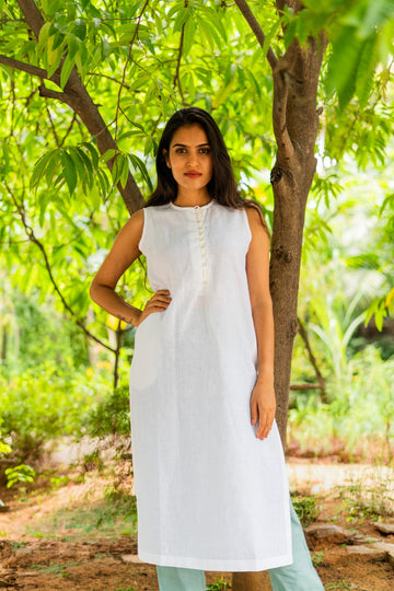 A woman wearing a sleeveless white kurta with a buttoned neckline, standing outdoors in front of a tree with green foliage.