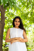 A woman with long dark hair wearing a sleeveless white top stands outdoors under a tree with green foliage in the background.