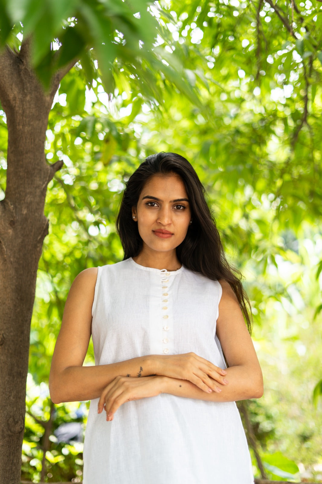 A woman with long dark hair wearing a sleeveless white top stands outdoors under a tree with green foliage in the background.