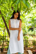 A woman wearing a sleeveless white kurta with a buttoned neckline, standing outdoors in a lush green garden setting.
