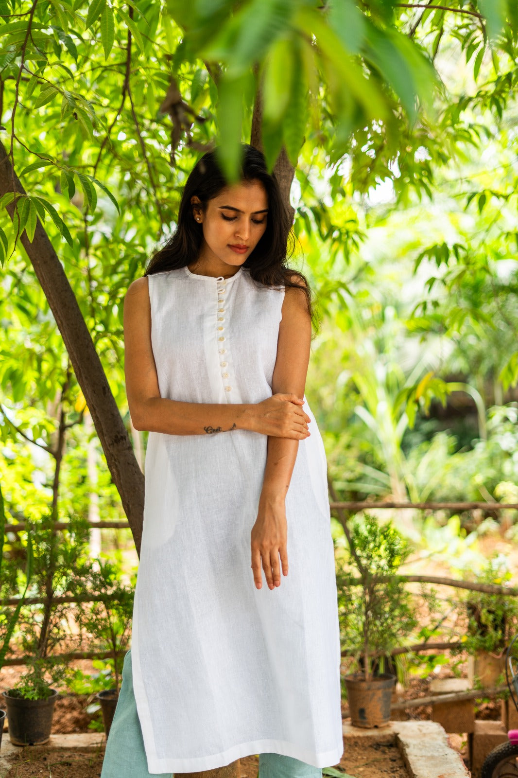 A woman wearing a sleeveless white kurta with a buttoned neckline, standing outdoors in a lush green garden setting.