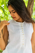 A woman wearing a sleeveless white linen top with a buttoned neckline, photographed outdoors against a soft-focus background of green leaves and tree trunks.
